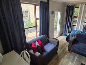 A cozy living area with a sofa, a chair, and a bed with a striped blanket. There is a sliding glass door leading to an outdoor area with plants.