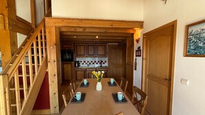 Een eetgedeelte met een lange tafel, stoelen, een vaas met bloemen en een keuken op de achtergrond.