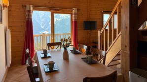Een houten chalet met een eettafel gedekt voor vier personen, een televisie aan de muur en uitzicht op besneeuwde bergen door grote ramen.