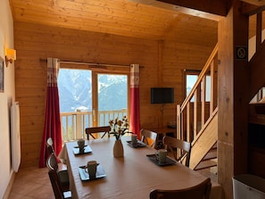 A wooden cabin with a dining table set for four, a television mounted on the wall, and a view of snow-capped mountains through large windows. - Spacious apartment, panoramic view - 6 guests (Villard-sur-Doron)