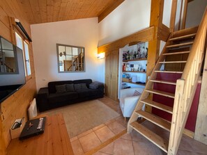 A cozy living room with a wooden ceiling, a sofa, a coffee table, a TV, a staircase, and a bookshelf. - Spacious apartment, panoramic view - 6 guests (Villard-sur-Doron)