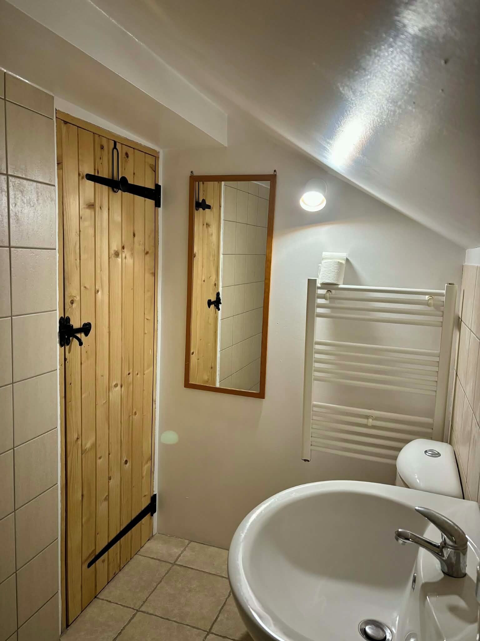 Une salle de bain avec une porte en bois, un miroir, un lavabo blanc et un porte-serviettes.