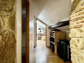 A narrow hallway with a stone wall on one side and a wooden door on the other. There is a wooden shelf unit with black luggage and a wooden beam overhead.