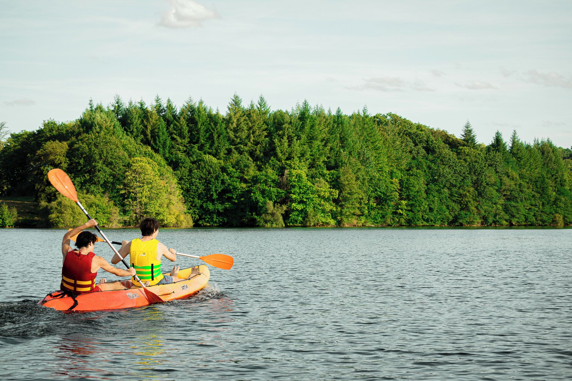 Two people kayaking on a calm lake with dense forest in the background.