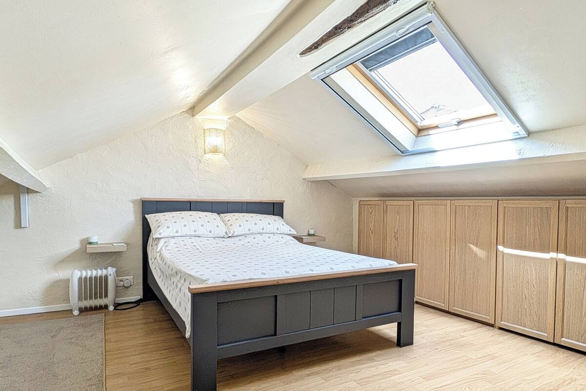 Un dormitorio con techo inclinado, tragaluz, cama con edredón estampado, radiador, suelo de madera y armarios empotrados de madera.