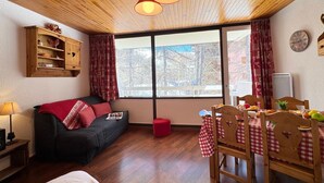 Et hyggeligt rum med træloft, en sofa, et spisebord med stole, et vindue med gardiner og et ur på væggen.