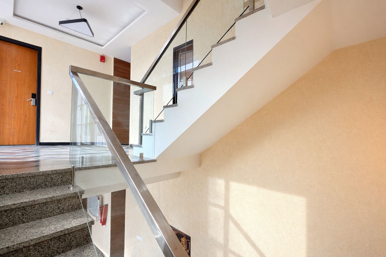 A modern interior with a glass staircase, a wooden door, and a ceiling light fixture.