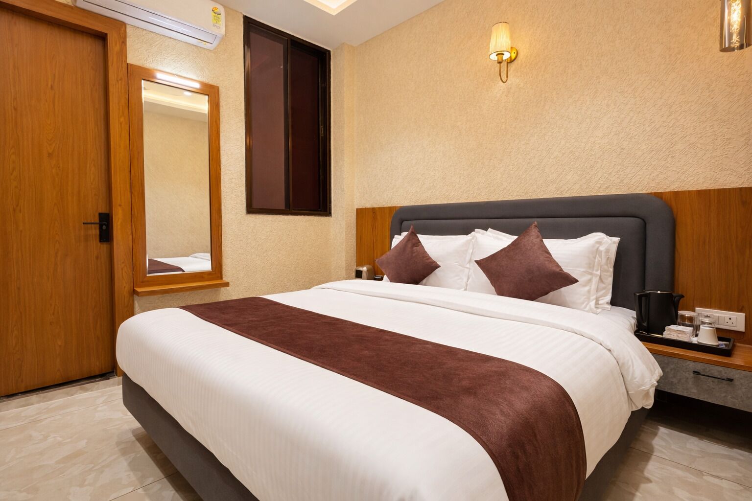 Une chambre d’hôtel comprenant un lit, une table de chevet, un miroir, une lampe fixée au mur et une porte.