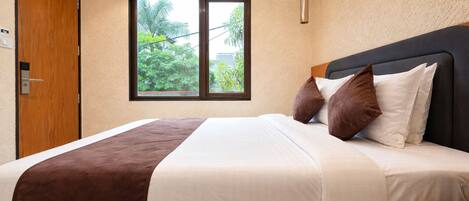 A hotel room with a bed, a window with a view of greenery, an air conditioning unit, and a bedside table.