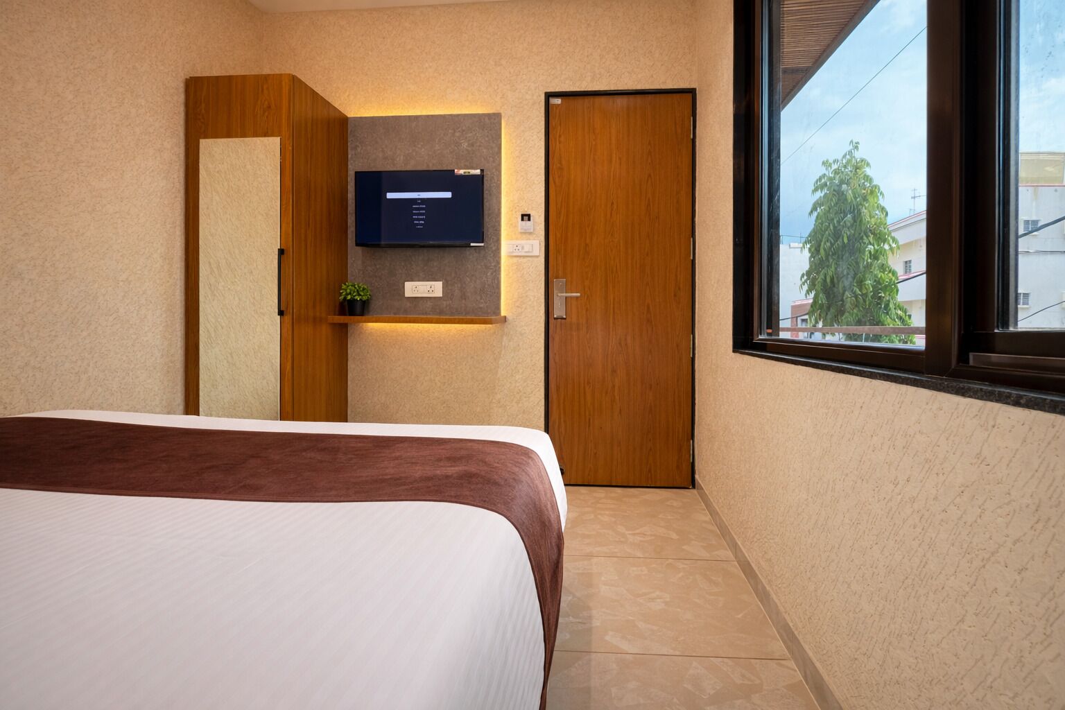 A hotel room with a bed, a TV mounted on the wall, a door, and a window with a view of buildings and trees.