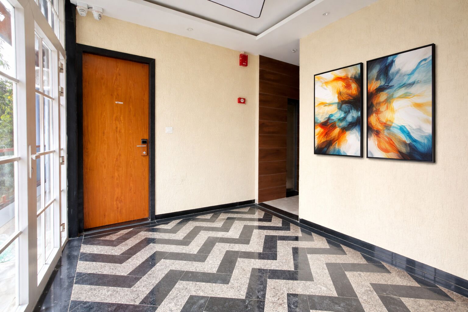 Un pasillo con una puerta de madera, un detector de incendios y dos cuadros abstractos en la pared.