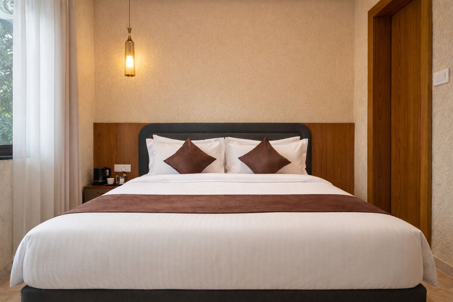 A hotel room with a large bed, a bedside table, a lamp, and a view of the outdoors.