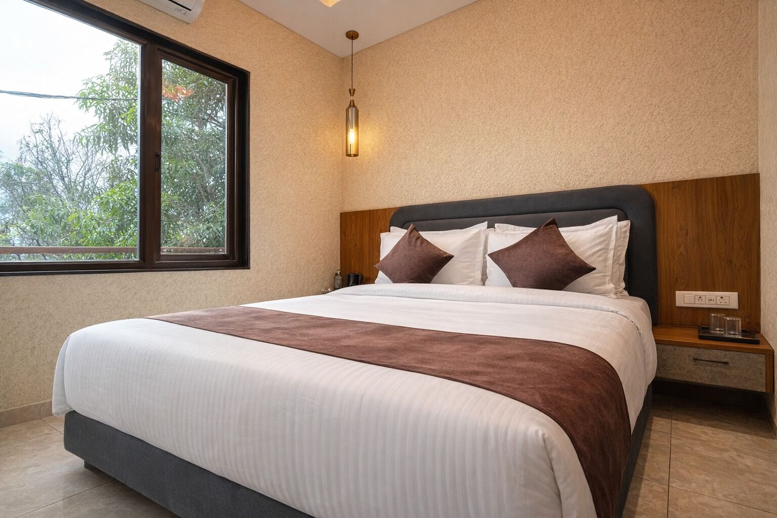 Una habitación de hotel con una cama grande, una mesita de noche, una lámpara de pared y una ventana con vistas a árboles.