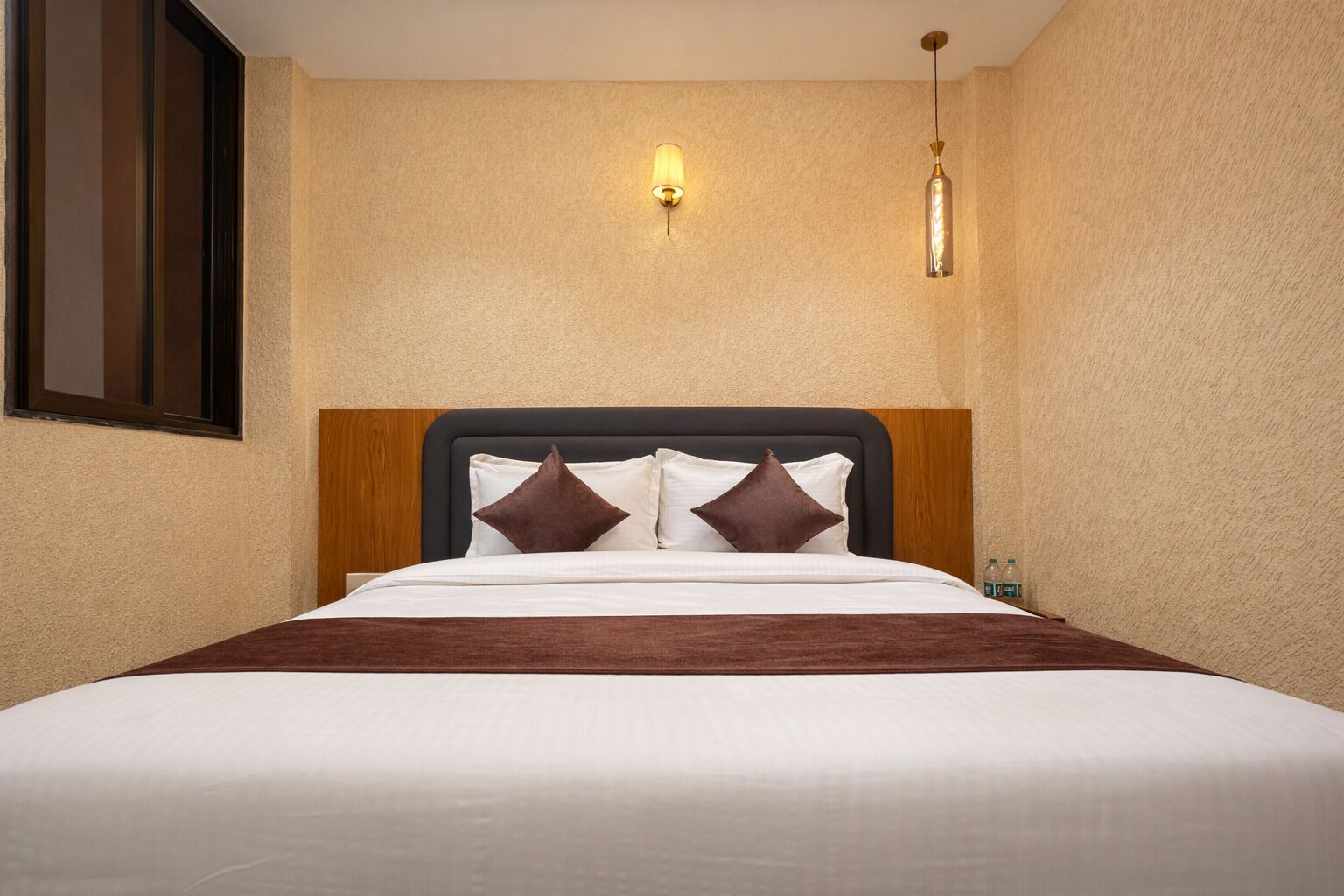 Habitación de hotel con cama, dos lámparas fijadas a la pared y cabecero de madera.