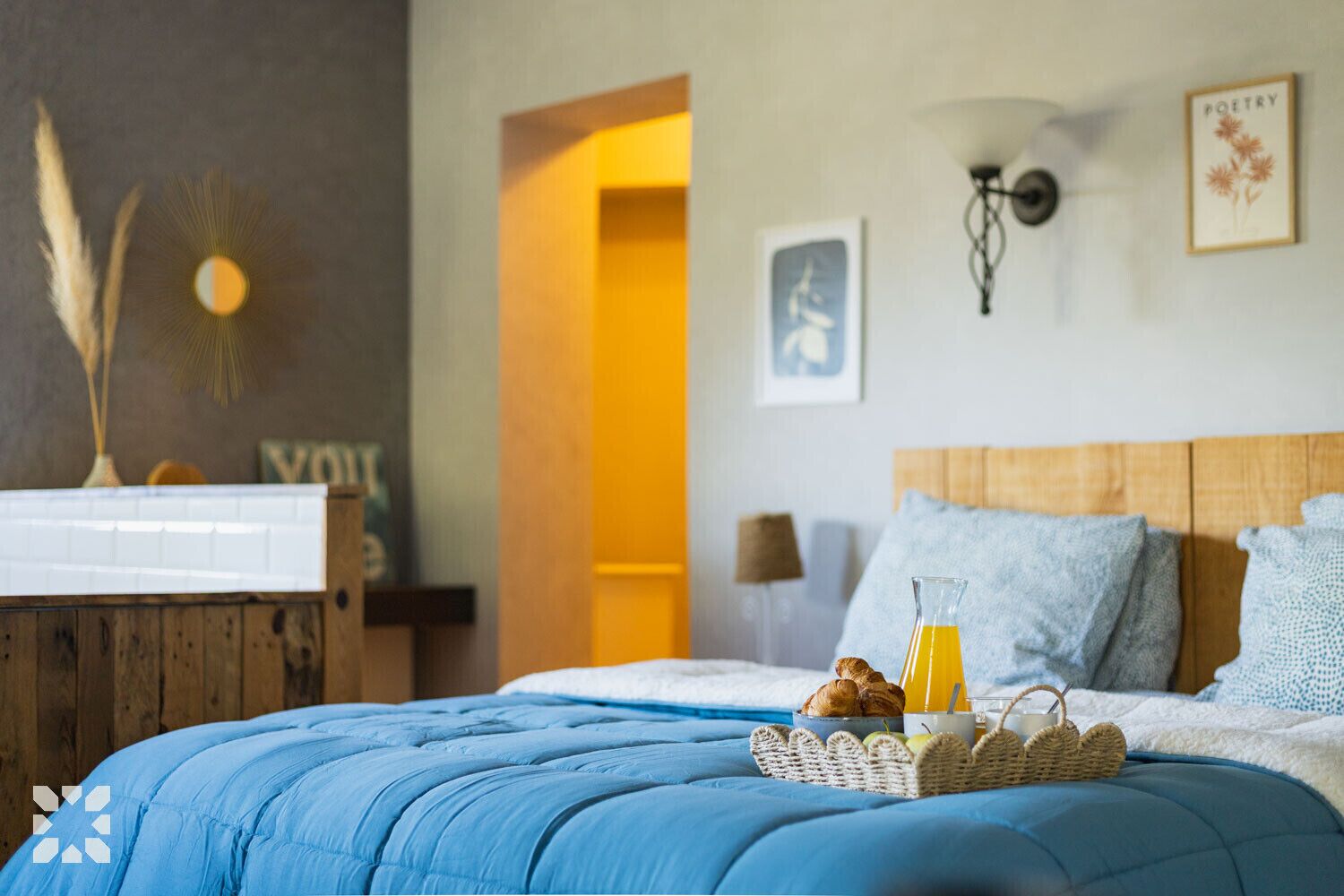 Un dormitorio con cabecera de madera, colcha azul, una cesta de pasteles y un vaso de jugo de naranja.