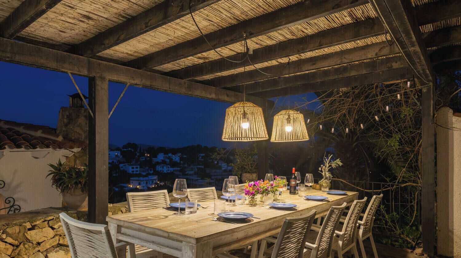 An outdoor dining area with a wooden table set for four, surrounded by wicker chairs, under a wooden pergola with hanging lights.