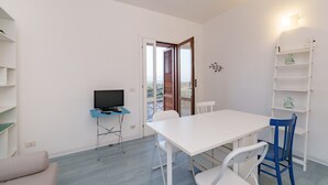 Ein Zimmer mit einem weißen Tisch, Stühlen, einem Fernseher auf einem Schrank, einem Bücherregal und einer Tür, die nach draußen führt.