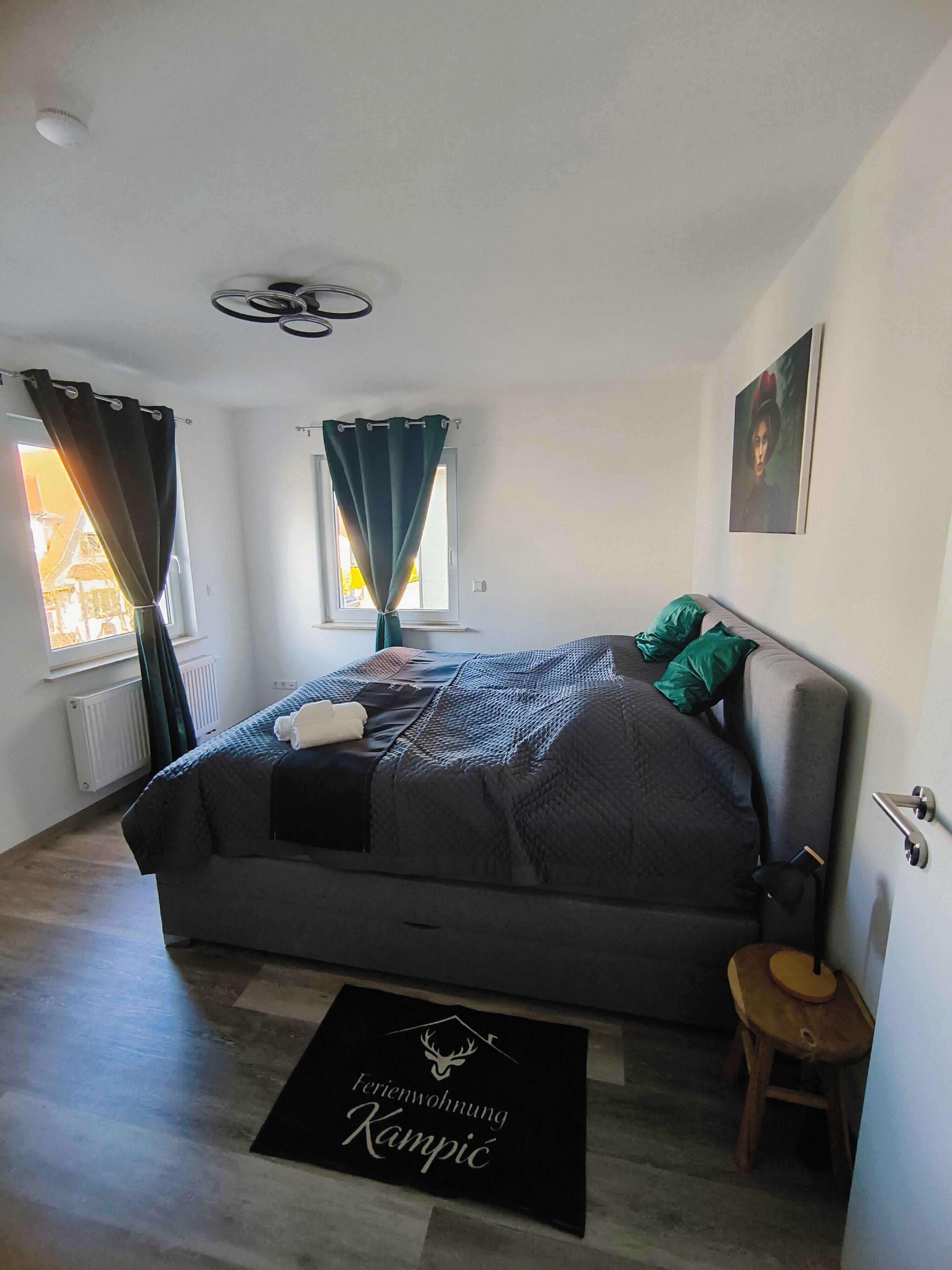 A bedroom with a bed, a bedside table, a window with curtains, a radiator, a door, and a doormat.