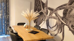 Un coin repas avec une table en bois, des chaises noires, un vase contenant des plumes d’herbe des pampas et une grande fresque murale représentant des bois de cerf.