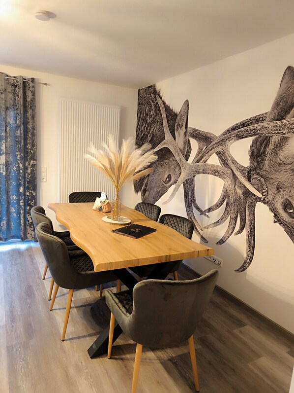 Un coin repas avec une table en bois, des chaises noires, un vase contenant des plumes d’herbe des pampas et une grande fresque murale représentant des bois de cerf.