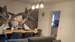 Un salon moderne avec un canapé d’angle, une table à manger avec des chaises, une grande fresque murale et une cuisine visible au fond.