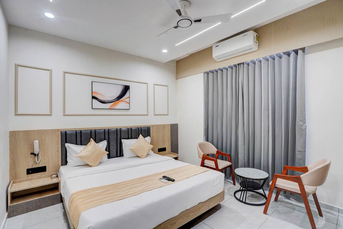 A modern hotel room with a large bed, a desk, two chairs, an air conditioning unit, and a ceiling fan.