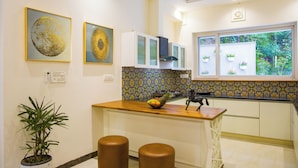 A kitchen with a fruit bowl, two stools, a potted plant, and a window with plants. - ELIVAAS Serenita Abode Tranquil Escape (Bagradhauran)
