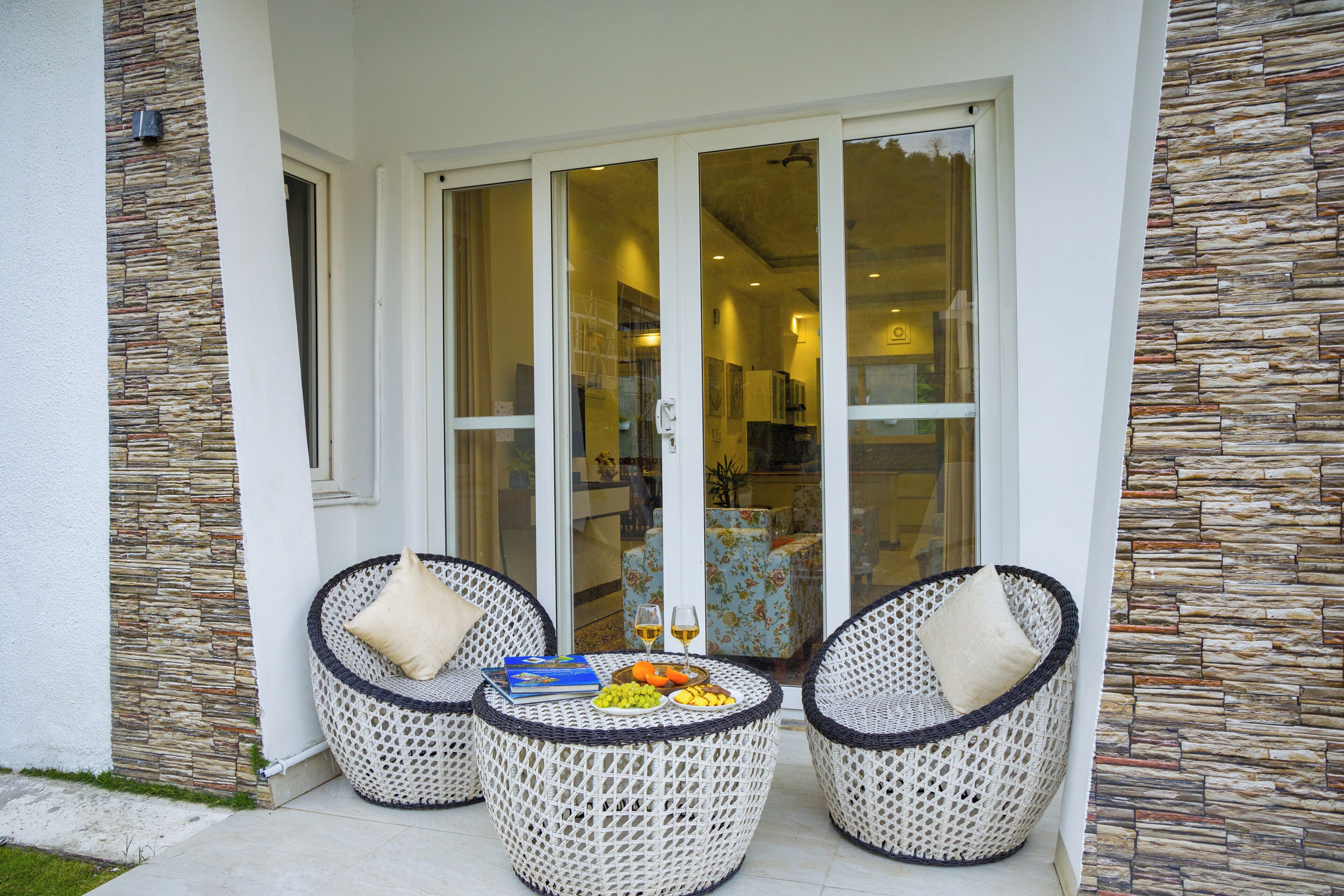 Un patio con mobiliario de mimbre, una mesa con frutas y vasos, y vistas a una habitación con pared de piedra a través de puertas de cristal.