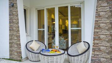 Un patio con mobiliario de mimbre, una mesa con frutas y vasos, y vistas a una habitación con pared de piedra a través de puertas de cristal.