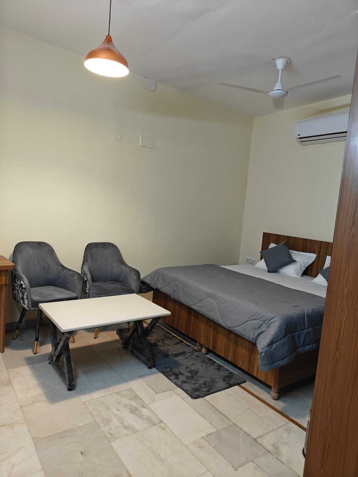A compact room with a bed, two chairs, a small table, a ceiling fan, and an air conditioning unit.