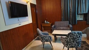 A modern living room with a flat-screen TV, wooden panel walls, a sofa, two patterned chairs, a coffee table, and a hanging pendant light. - The Grey Leaf Residency (New Delhi)