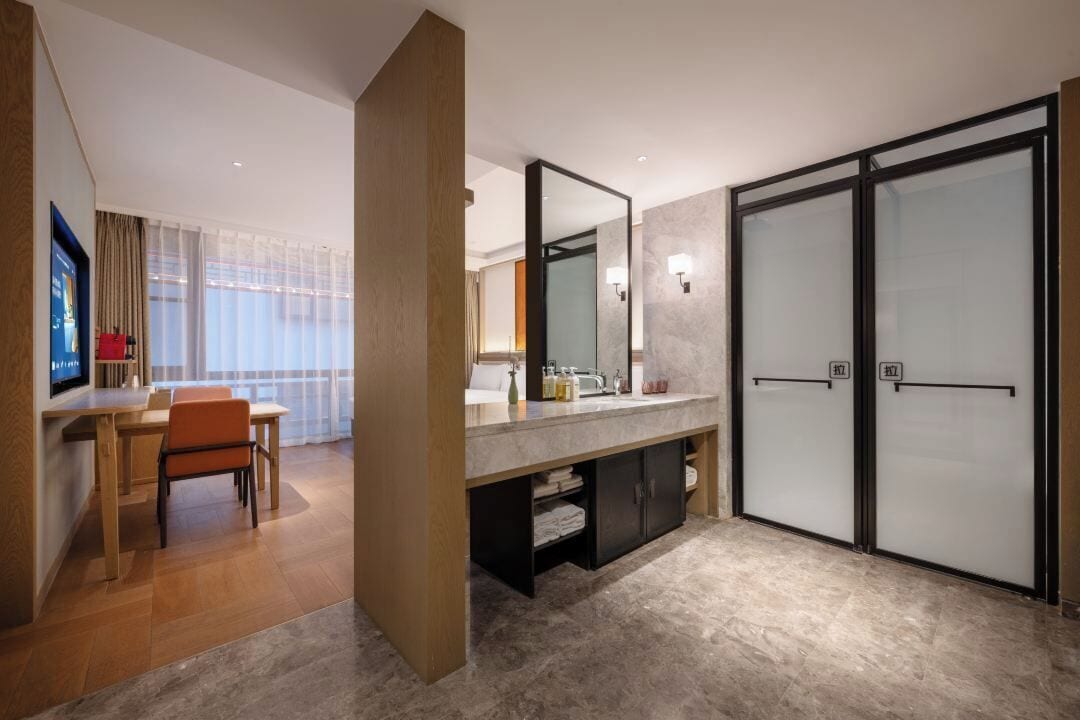 A modern hotel room with a wooden desk, a large mirror, a bathroom with frosted glass doors, and a television on the left.