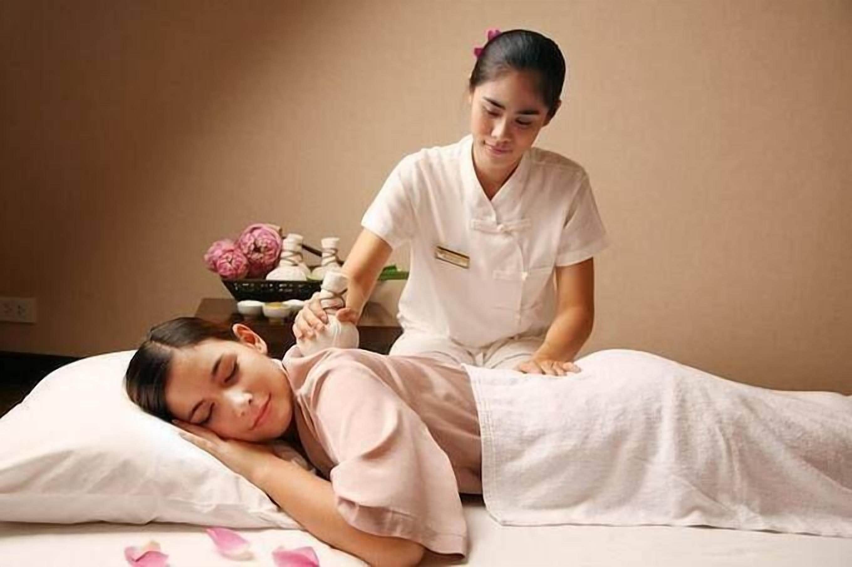 A person lying on a bed receiving a massage, with a massage table and a small table with flowers and a bowl in the background.
