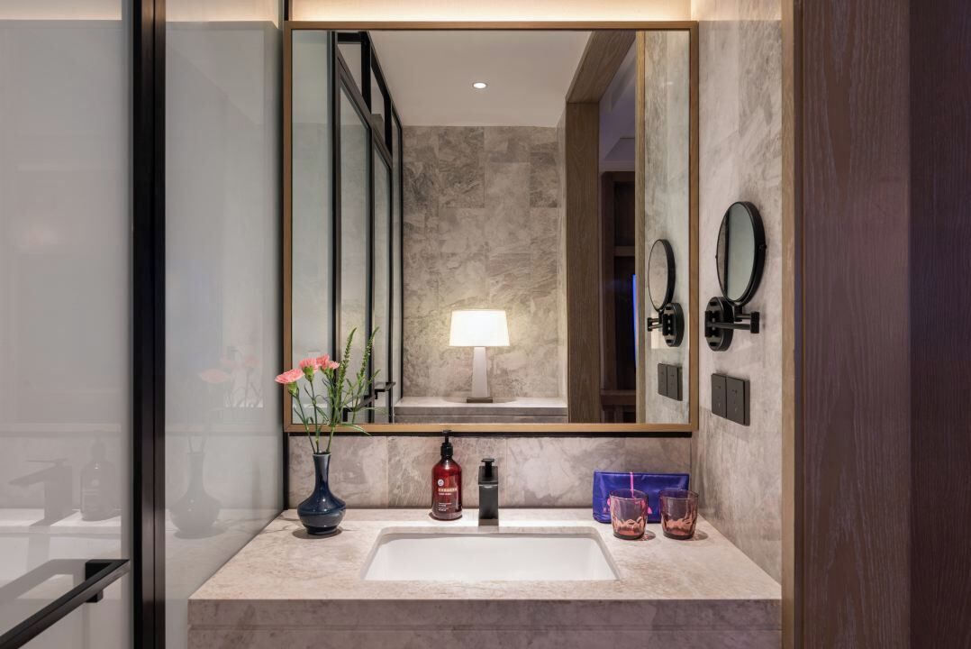 Een badkamer met een grote spiegel, een wastafel, een vaas met bloemen, een zeepfles en een glas water.