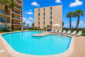 A swimming pool with lounge chairs, palm trees, and a building with balconies. - Marks Beachfront Condo with 3rd Floor Balcony | Amazing Views + Pool (South Padre Island)