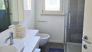 Casa de banho com lavatório branco, espelho e sanita, zona de duche e uma pequena janela.