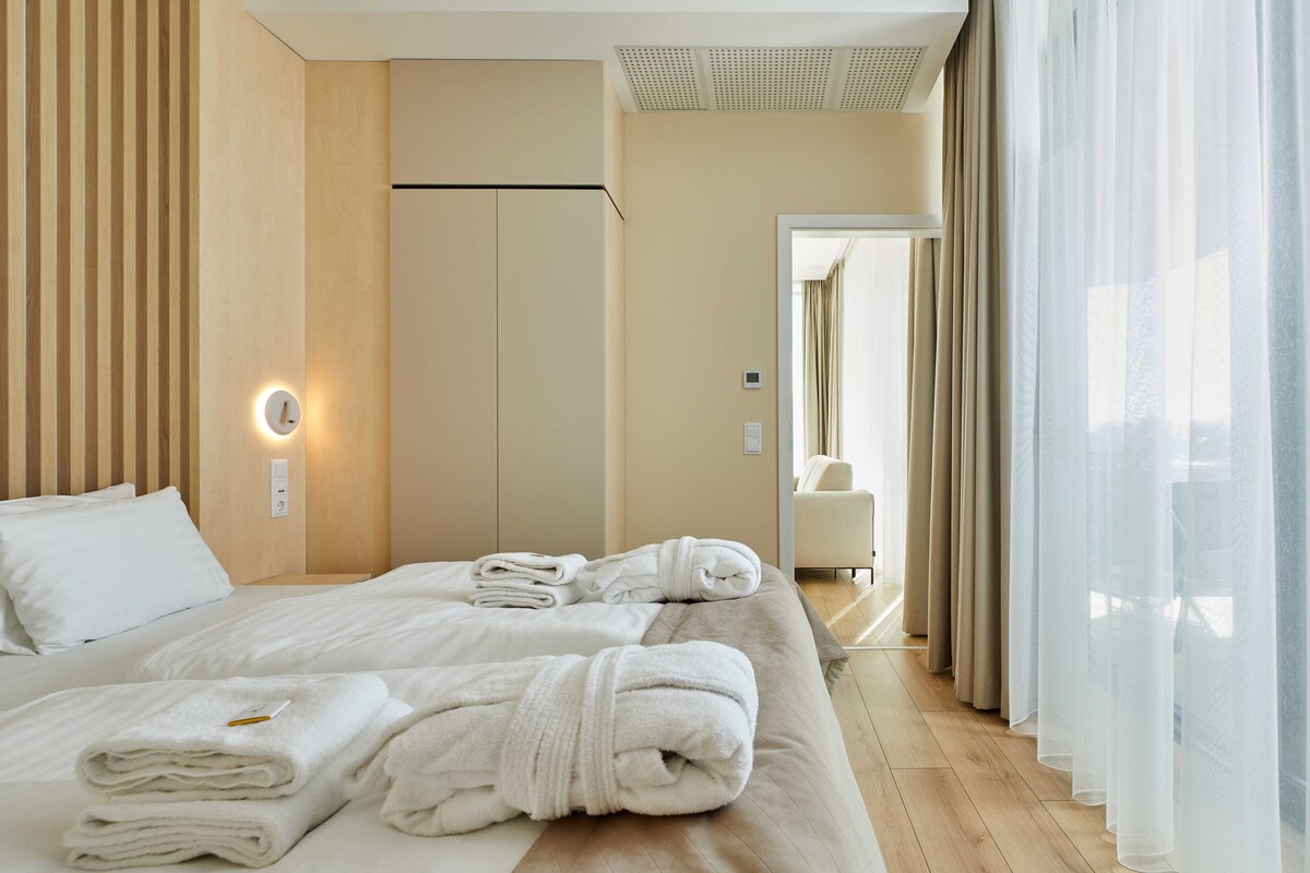 A hotel room with a bed, towels, a nightstand, a wall clock, and a window with curtains.
