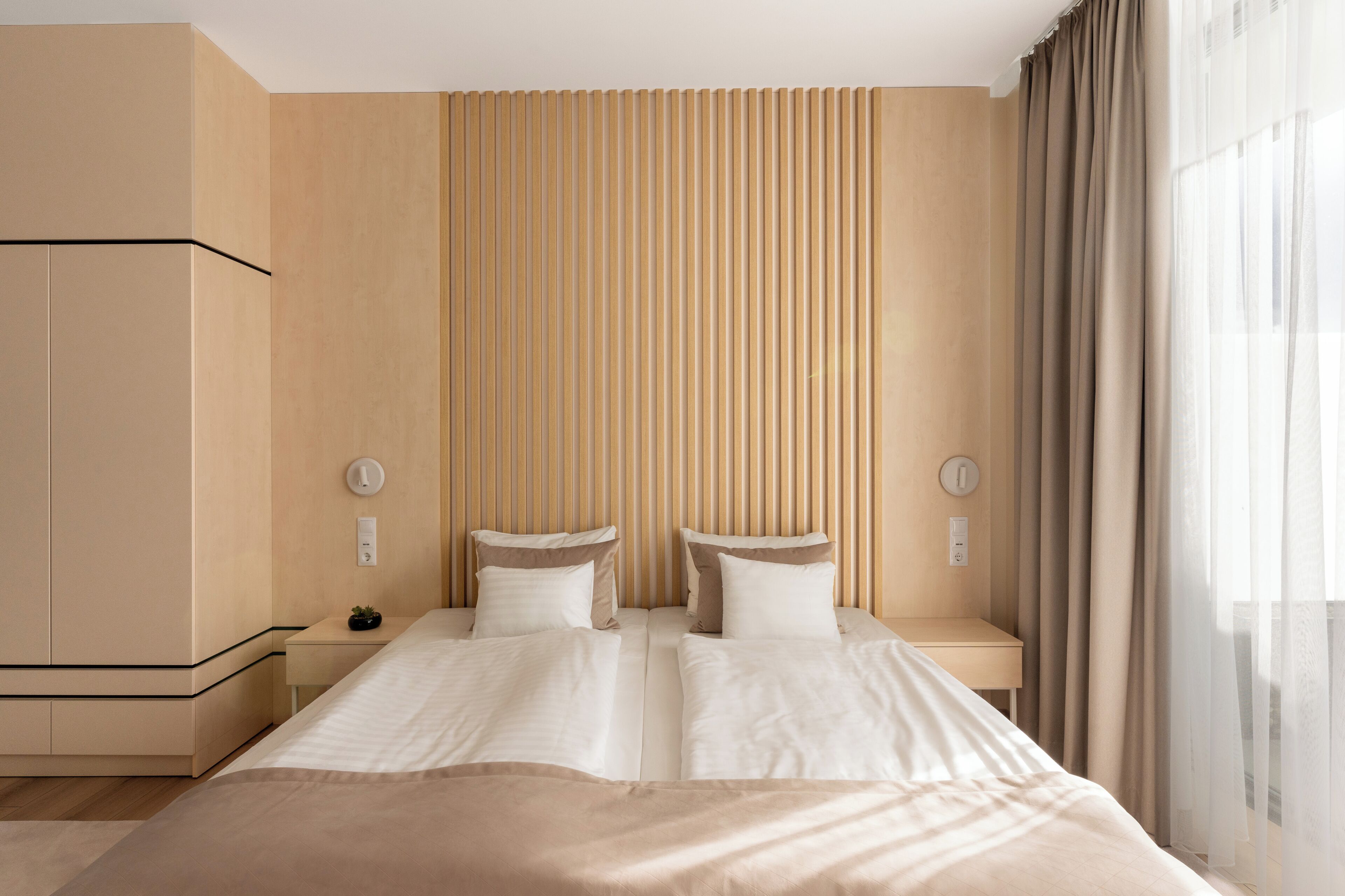 A double bed with two pillows, a bedside table with a lamp, a wall-mounted clock, and a window with curtains.