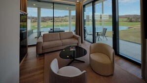A modern living room with a sofa, armchair, and coffee table, large windows with a view of a golf course and lake, and a flat-screen TV mounted on the wall. - Evergreen Health (Kehidakustány)