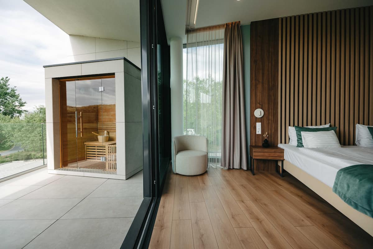 A modern bedroom with a large bed, a seating area, and a sliding glass door leading to a balcony.