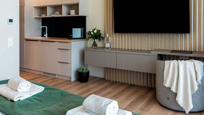 A modern hotel room with a wooden floor, a green bed, a grey sofa, a flat-screen TV, a small kitchen area, and a potted plant. - Evergreen Health (Kehidakustány)