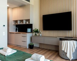 Una habitación de hotel moderna con piso de madera, una cama verde, un sofá gris, un televisor de pantalla plana, una pequeña área de cocina y una planta en maceta.