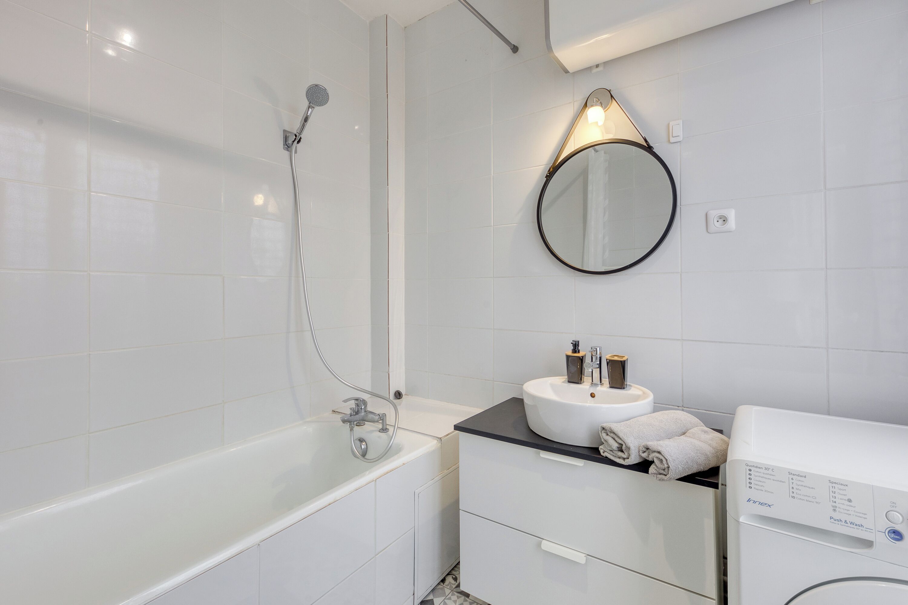 Une salle de bain moderne avec une baignoire, un miroir rond, un lavabo avec une serviette et un lave-linge.