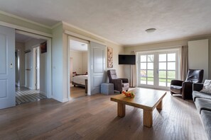 A spacious living room with hardwood flooring, a wooden coffee table, a sofa, a recliner, and a television mounted on the wall.