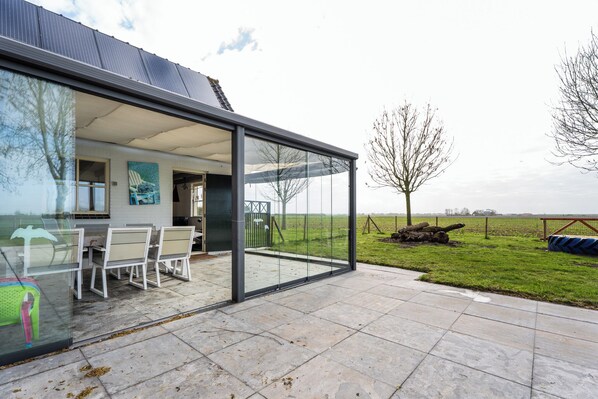 A modern house with a glass-enclosed patio, a dining area with chairs, and a view of a grassy field.