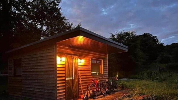 A small wooden cabin with a porch, a bicycle, and a fire pit. - Craigengillan Mini Lodge (Dalmellington)