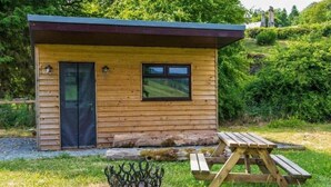 A small wooden cabin with a door, window, and picnic table in a grassy area. - Craigengillan Mini Lodge (Dalmellington)