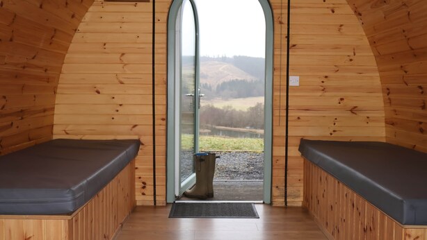 A wooden interior with a large arched doorway, two benches, and a view of a landscape. - Accessibile Craigengillan Cabin (Dalmellington)