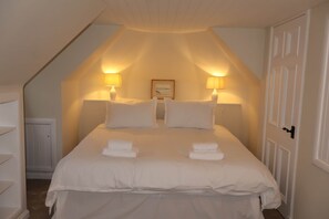 Ein Schlafzimmer mit schräg verlegtem Dach, zwei Nachttischlampen, einem gerahmten Bild an der Wand und ordentlich gefalteten Handtüchern auf dem Bett.