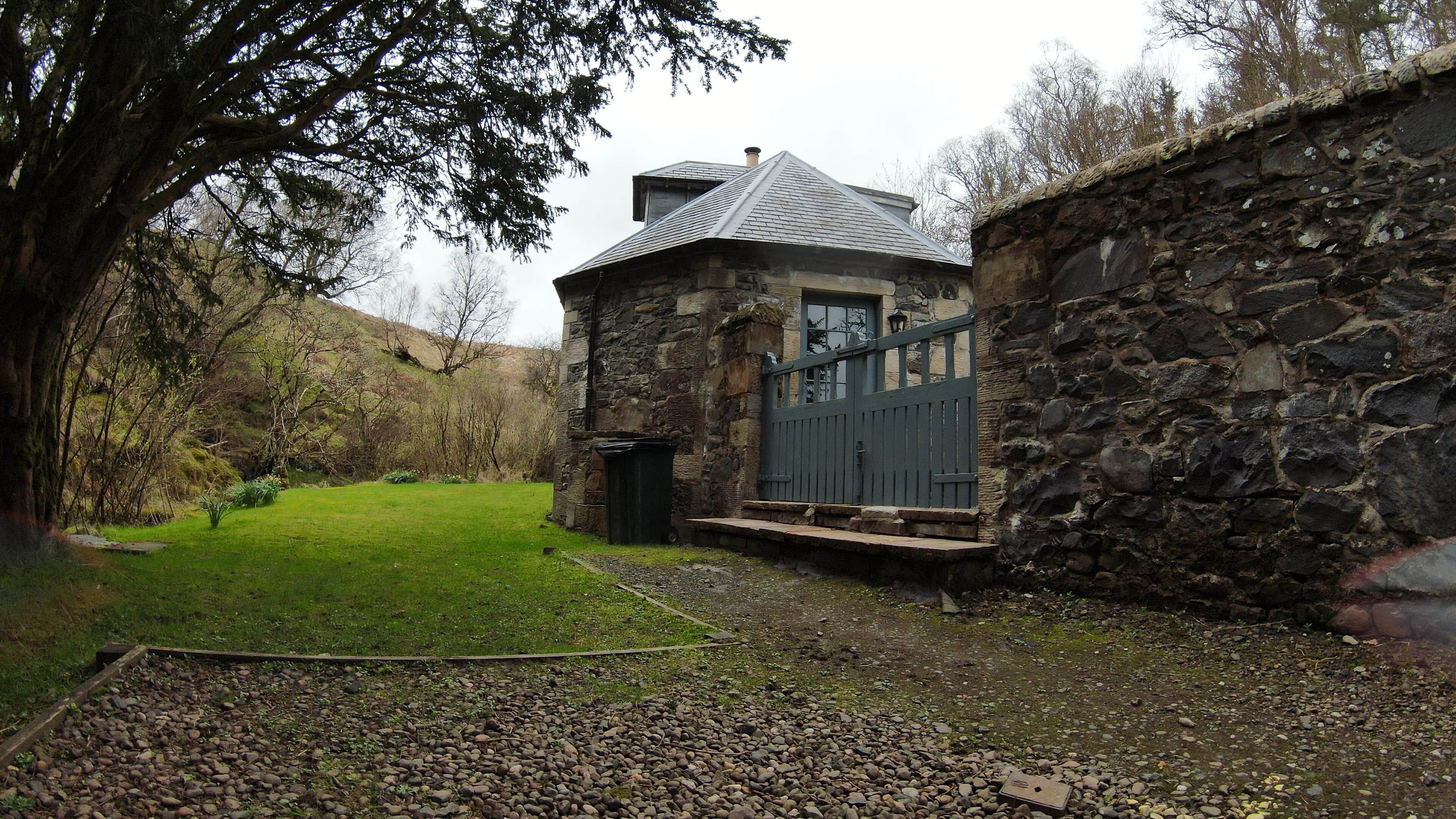 A stone building with a metal roof, a paved area with pebbles, and a grassy yard.